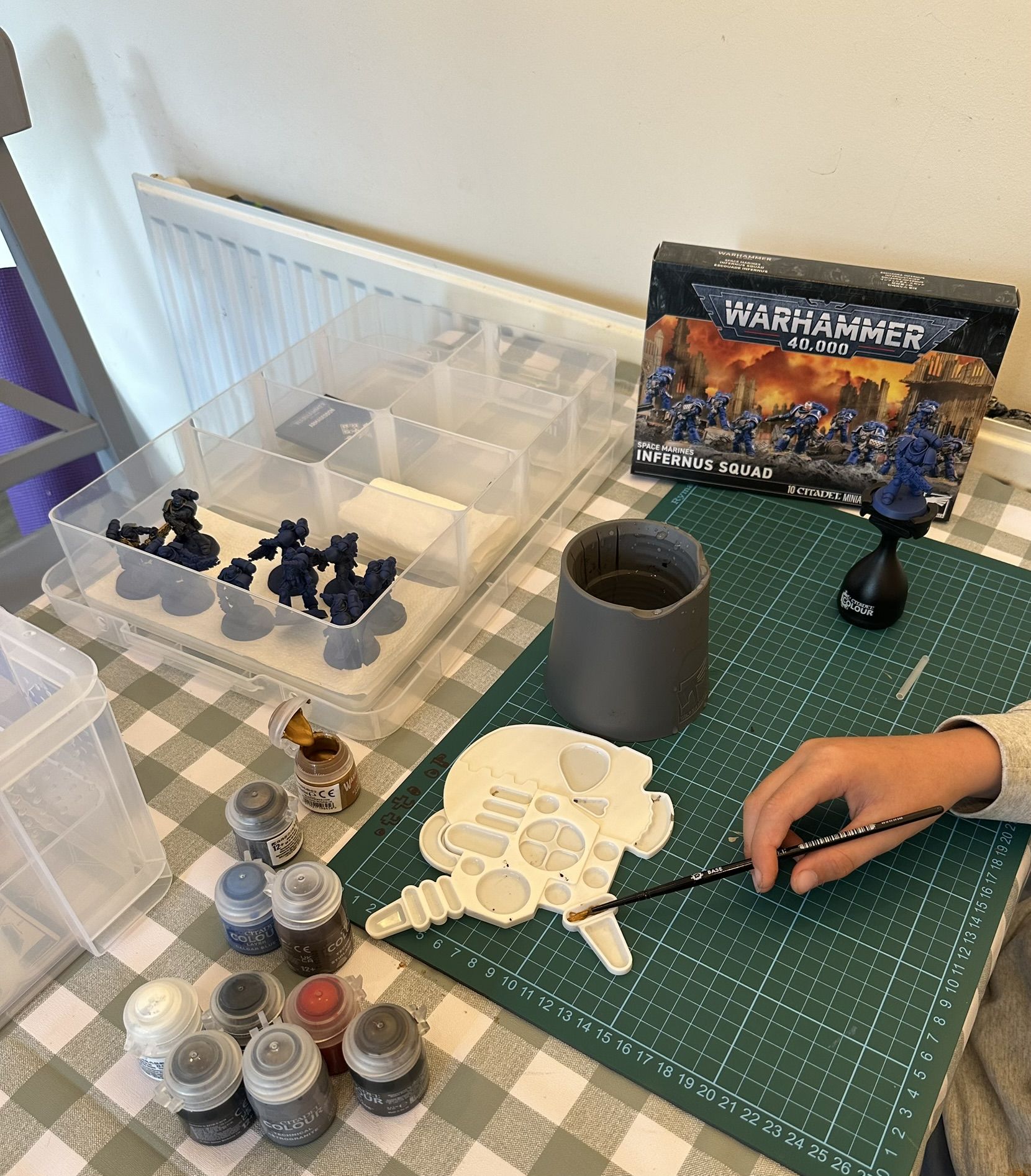 A painting table mid session, brush in hand over a palette, Citadel paints, a water pot, the Infernus Squad box and a tray of blue primed Space Marines.