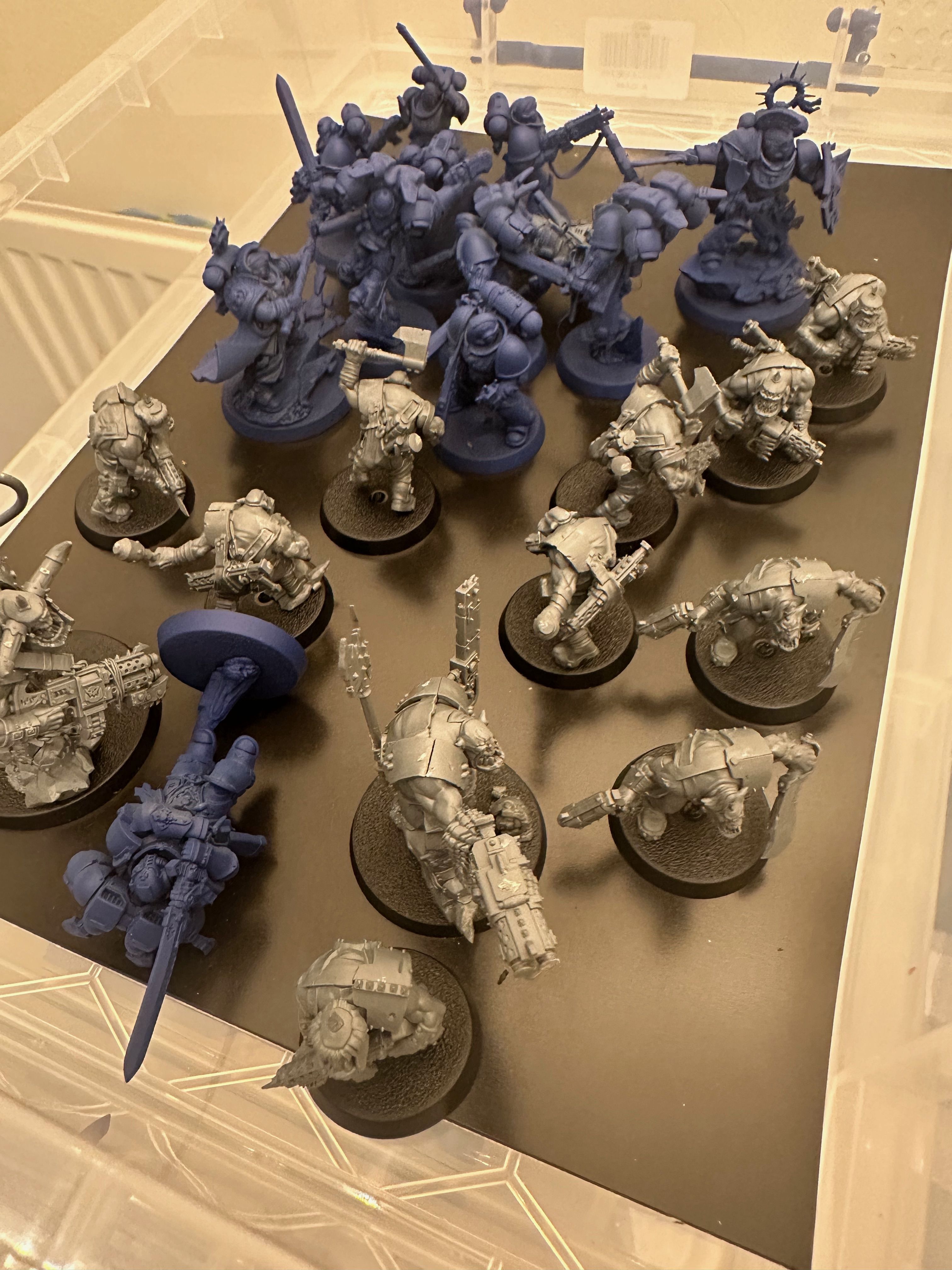 An open clear plastic storage box lined with dark card, full of unfinished Warhammer miniatures. Blue-primed Space Marines crowd the back, bare grey Orks on black bases fill the front, and one Marine lies toppled on its side.